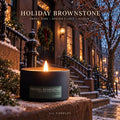 Holiday Brownstone pine and spiced clove candle burning in a matte black tin on a snowy Brooklyn brownstone stoop