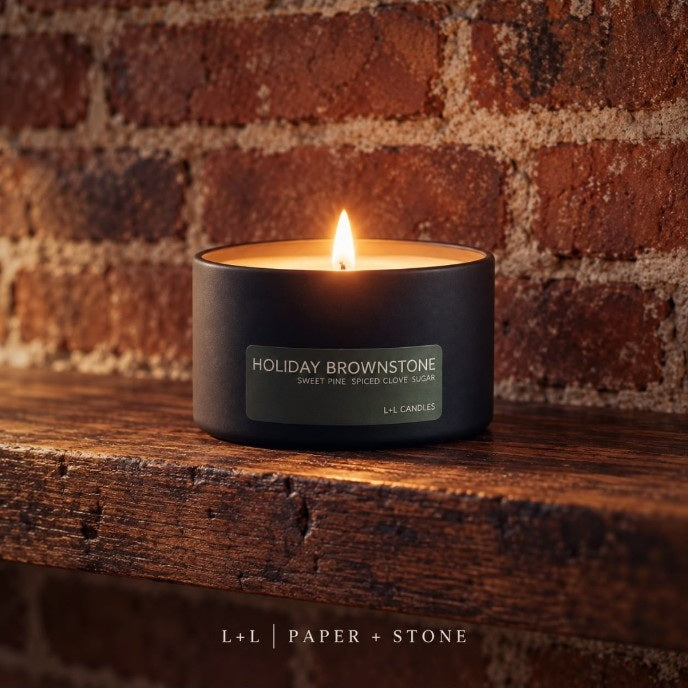 Lit Holiday Brownstone candle in a black travel tin on a wooden shelf against a warm rustic brick wall
