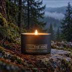 The Hike pine, fir and cedar scented candle in a matte black tin, lit on a mossy stone against a misty evergreen forest
