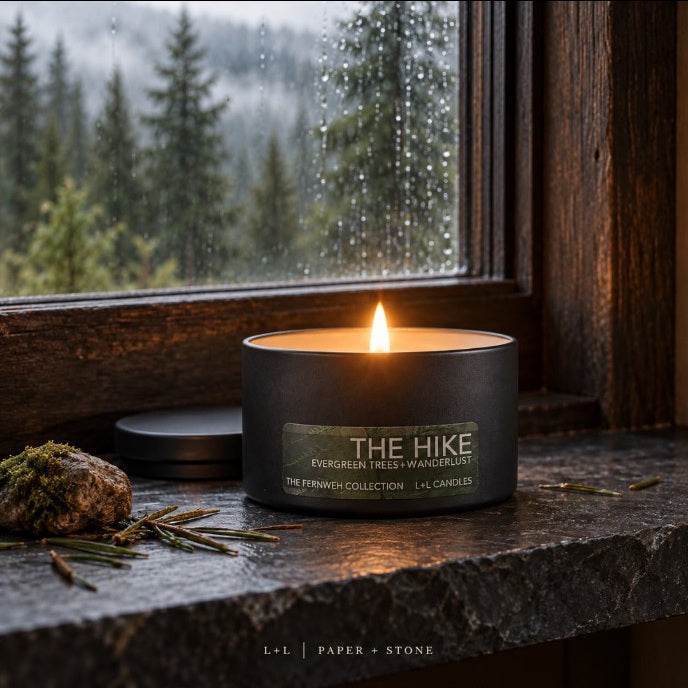 The Hike woodland scented candle burning on a rustic stone windowsill beside a rain-speckled forest view