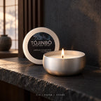 Tōjinbō sea-salt scented candle burning in a brushed silver tin beside its labeled lid on a stone ledge